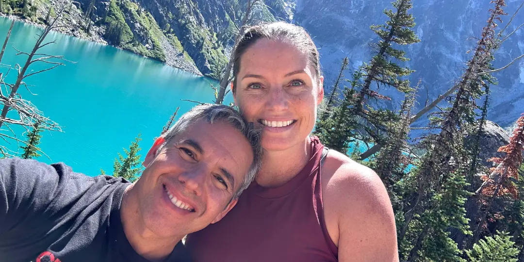 Casti and Juan Cava, owners of Casti Buys Houses, smiling together on a hike with a beautiful mountain lake in the background, representing their personal, hands-on approach to home buying.